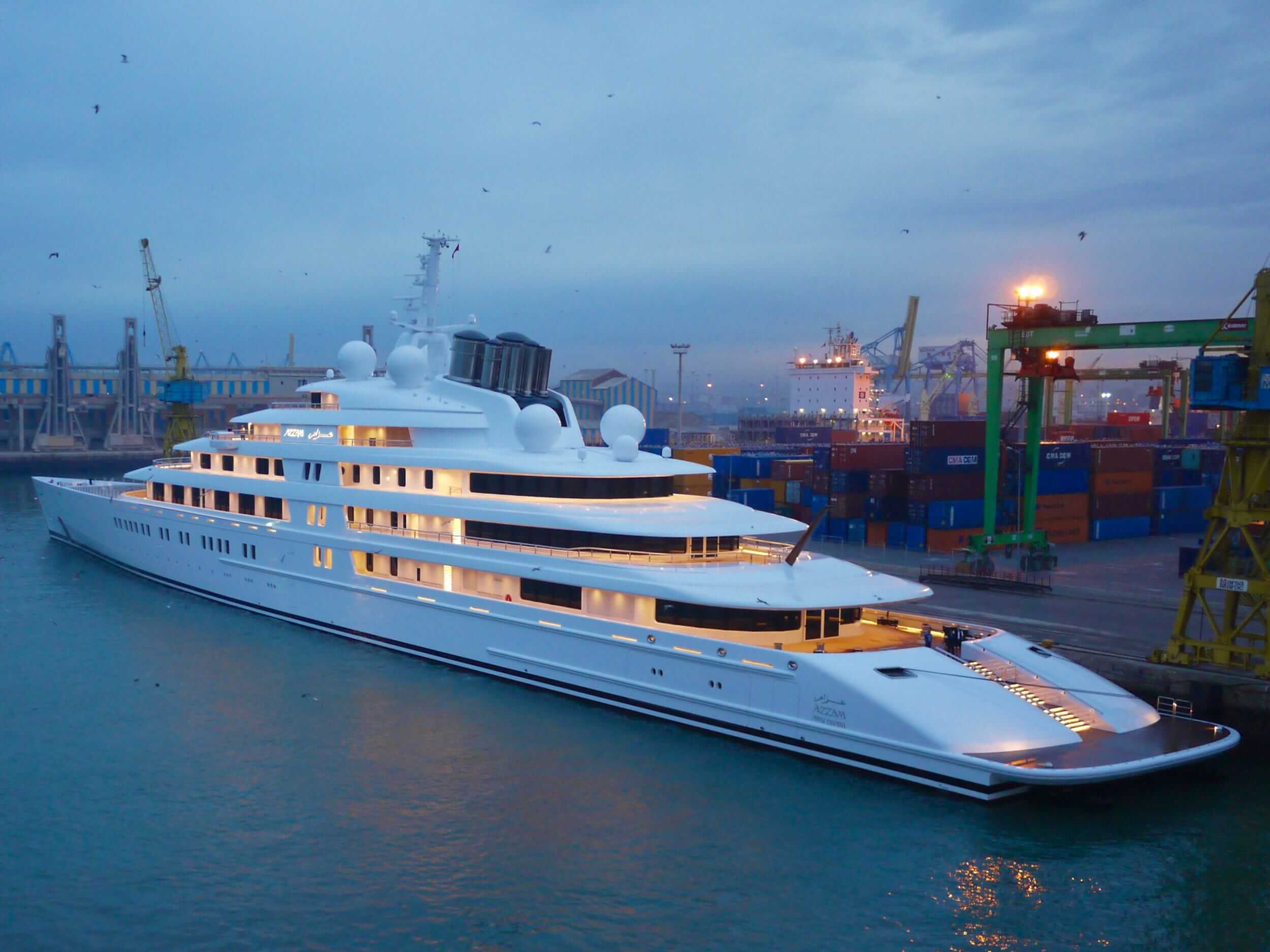 Luxury superyacht Azzam docked at port, showcasing sleek white design and illuminated decks, surrounded by shipping containers and cranes.