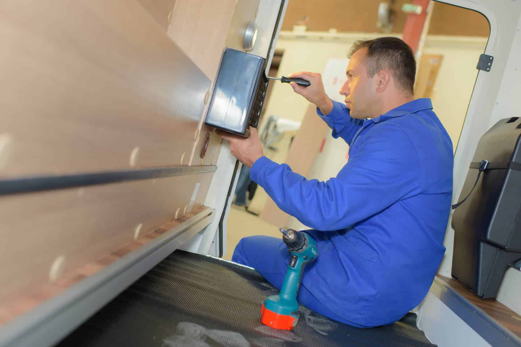 Technician installing marine electrical panel in yacht, using screwdriver and power drill, surrounded by wooden interior.