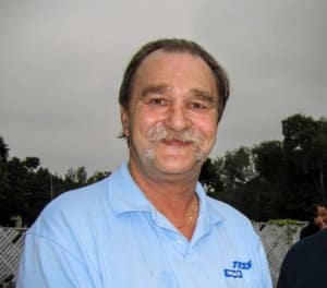 Smiling man wearing a light blue Tess Electrical LLC shirt, standing outdoors with trees in the background, in memory of Frank Salenieks.