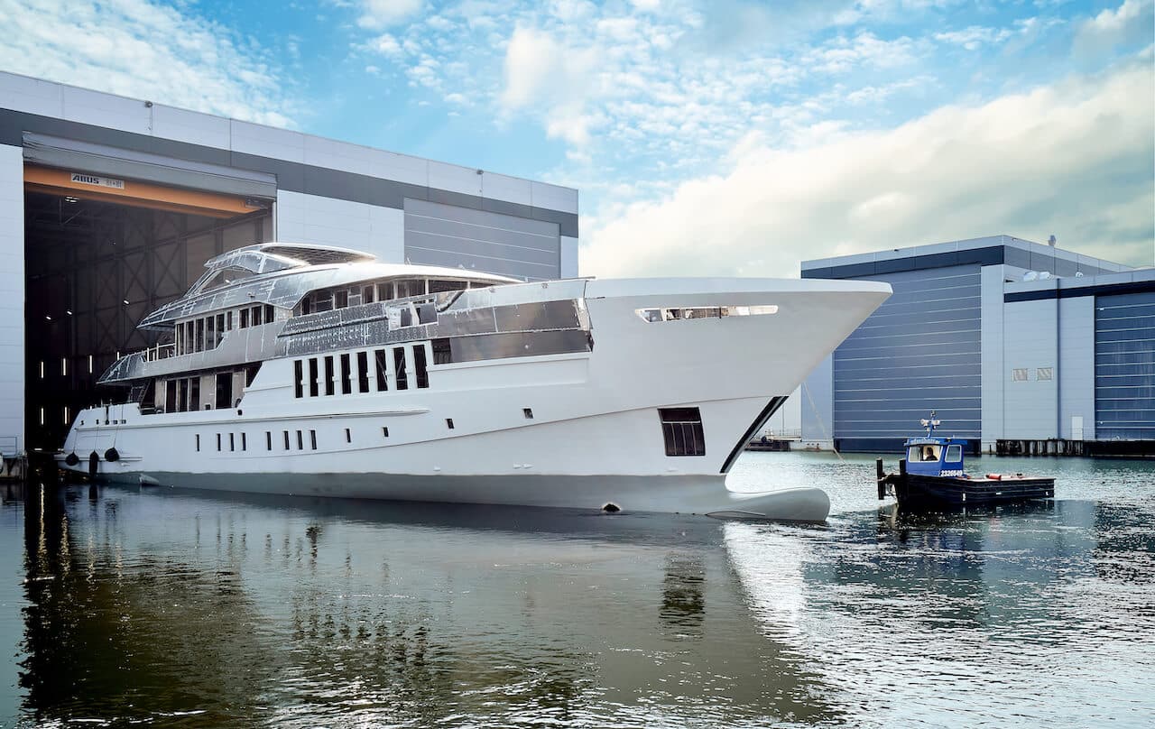 Luxury yacht Project Pollux emerging from the Heesen shipyard, showcasing modern design elements and sleek lines, with a small support boat nearby and industrial buildings in the background.