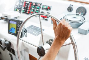 Person steering a boat with a ship's wheel and electronic navigation equipment, highlighting marine electrical systems and control interfaces relevant to Tess Electrical's services.