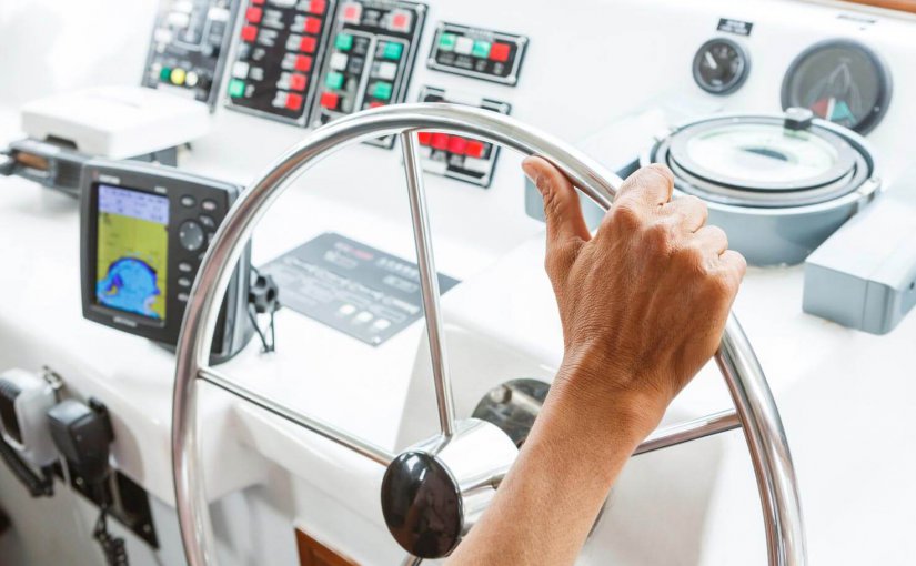 Hand gripping yacht steering wheel with navigation instruments and control panel in the background, illustrating marine electrical systems and operational safety.