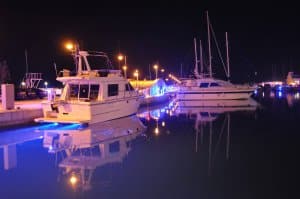 Boats docked at night illuminated by blue and yellow dock lights, reflecting in calm water, showcasing proper lighting for safe marine environments.