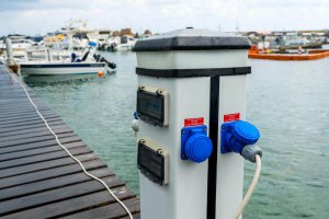 Marina power pedestal with blue electrical outlets and meters, surrounded by boats on water, illustrating essential marina electrical equipment for safe operations.