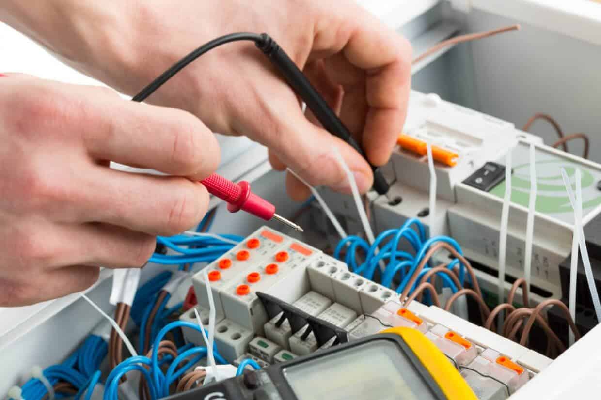 Hands using multimeter probes on electrical wiring and components within a residential electrical panel, illustrating electrical contracting services for upgrades and installations.