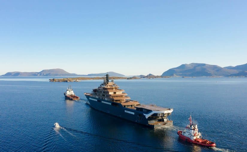 REV Ocean, the largest yacht in the world, being towed in open water with tugboats, showcasing its impressive size and design against a scenic backdrop of mountains and clear skies.