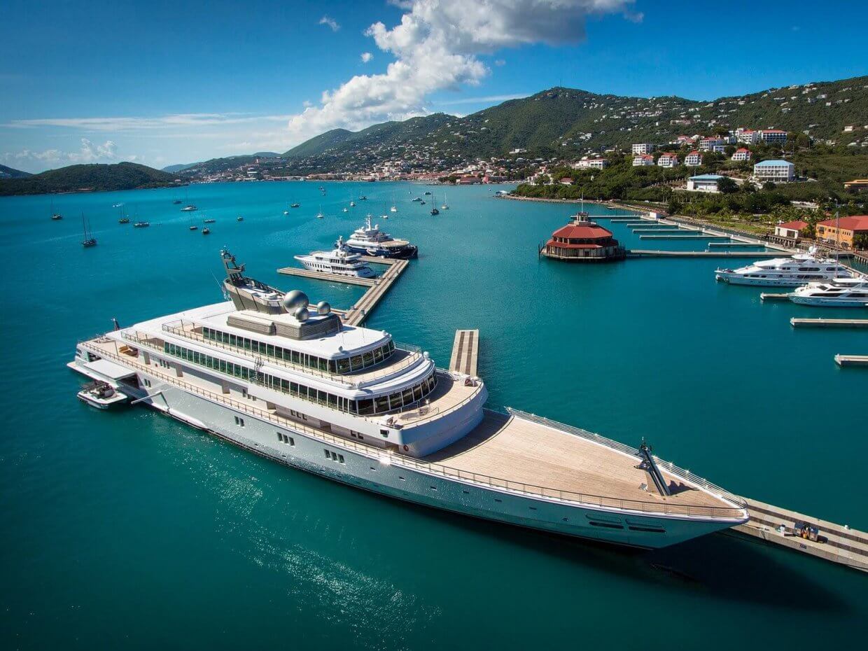 Mega yacht docked in a vibrant marina, showcasing marine engineering and design, surrounded by commercial vessels and scenic hills.