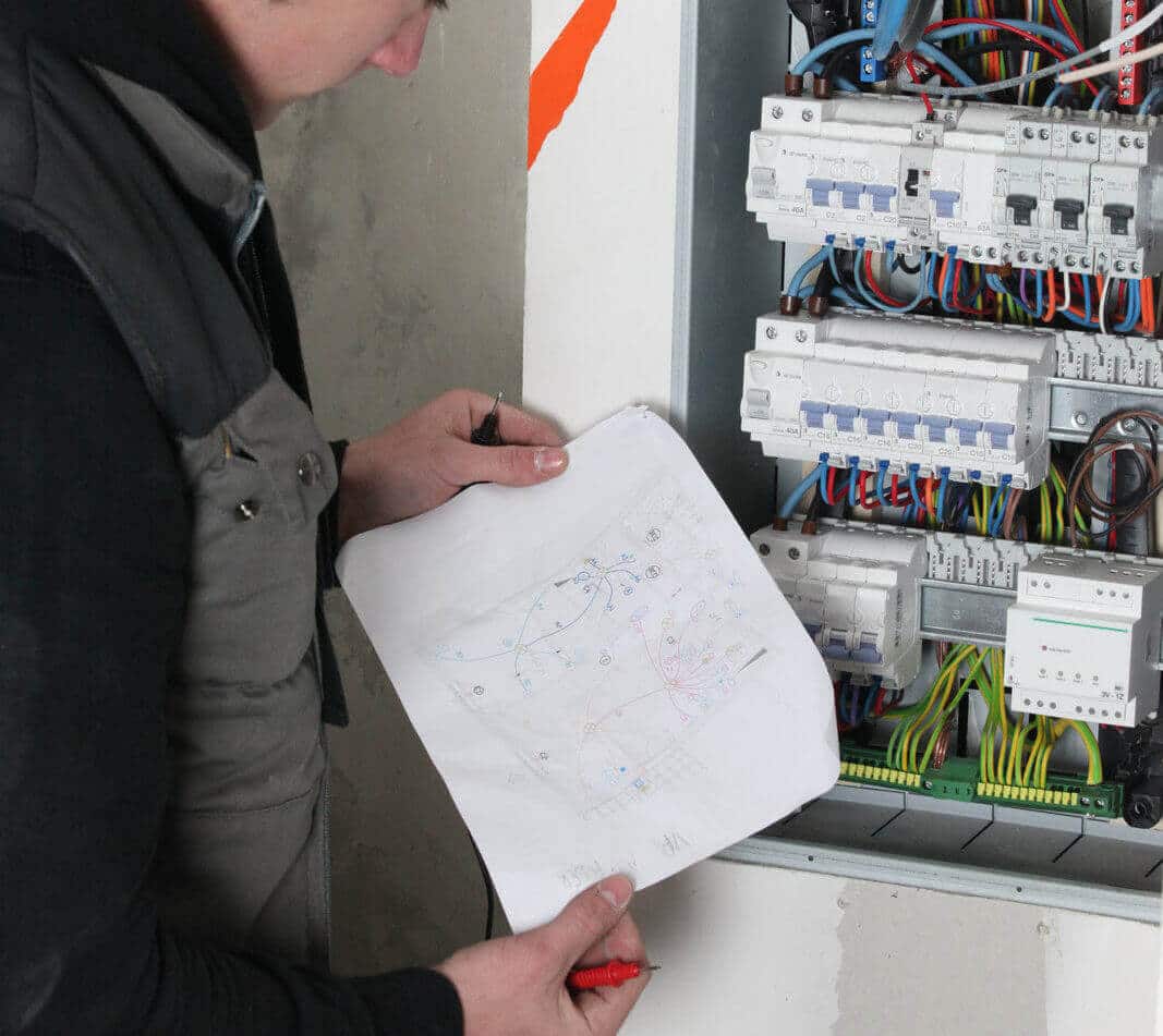 Electrician analyzing wiring diagram next to electrical panel, illustrating yacht electrical system maintenance and fault finding services.