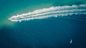 Aerial view of a powerboat creating a wake in the ocean, with a sailboat in the distance, highlighting marine activities and yachting culture.