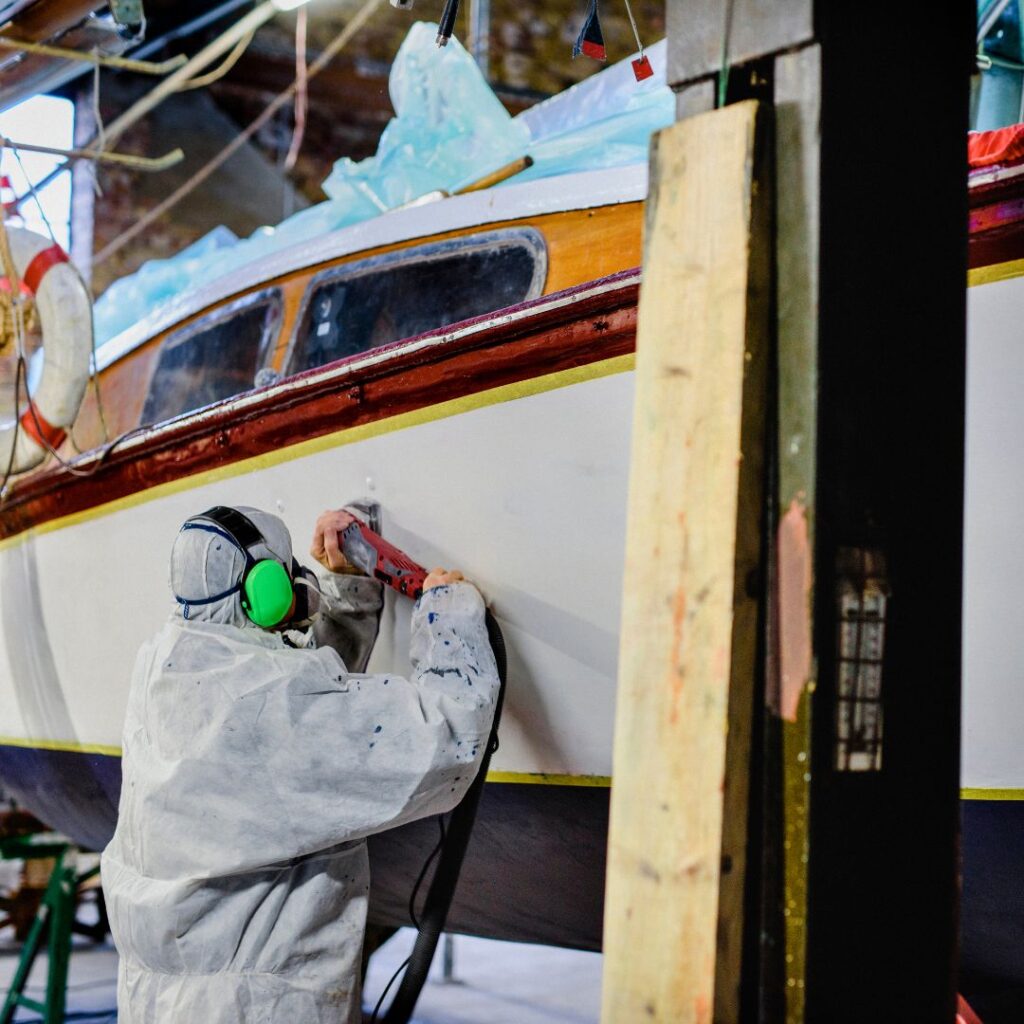 Person in protective gear sanding the hull of a yacht in a marine maintenance facility, emphasizing yacht maintenance and repair services.