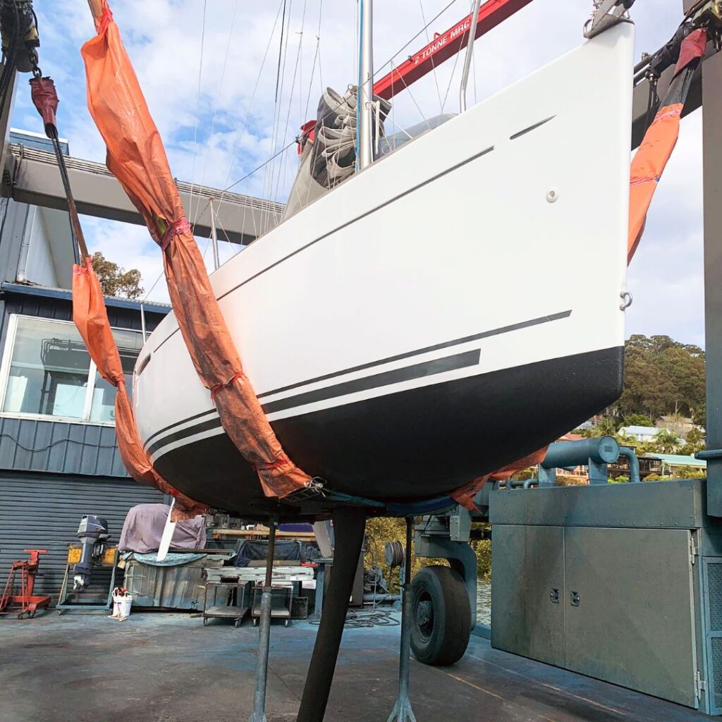 Yacht being lifted for marine electrical refit, showcasing orange lifting straps and surrounding maintenance equipment.