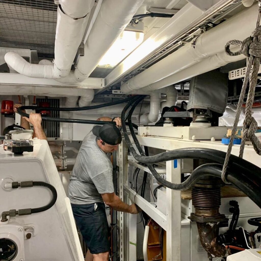 Technician performing maintenance on yacht's power system, inspecting cables and components in a marine engine room.
