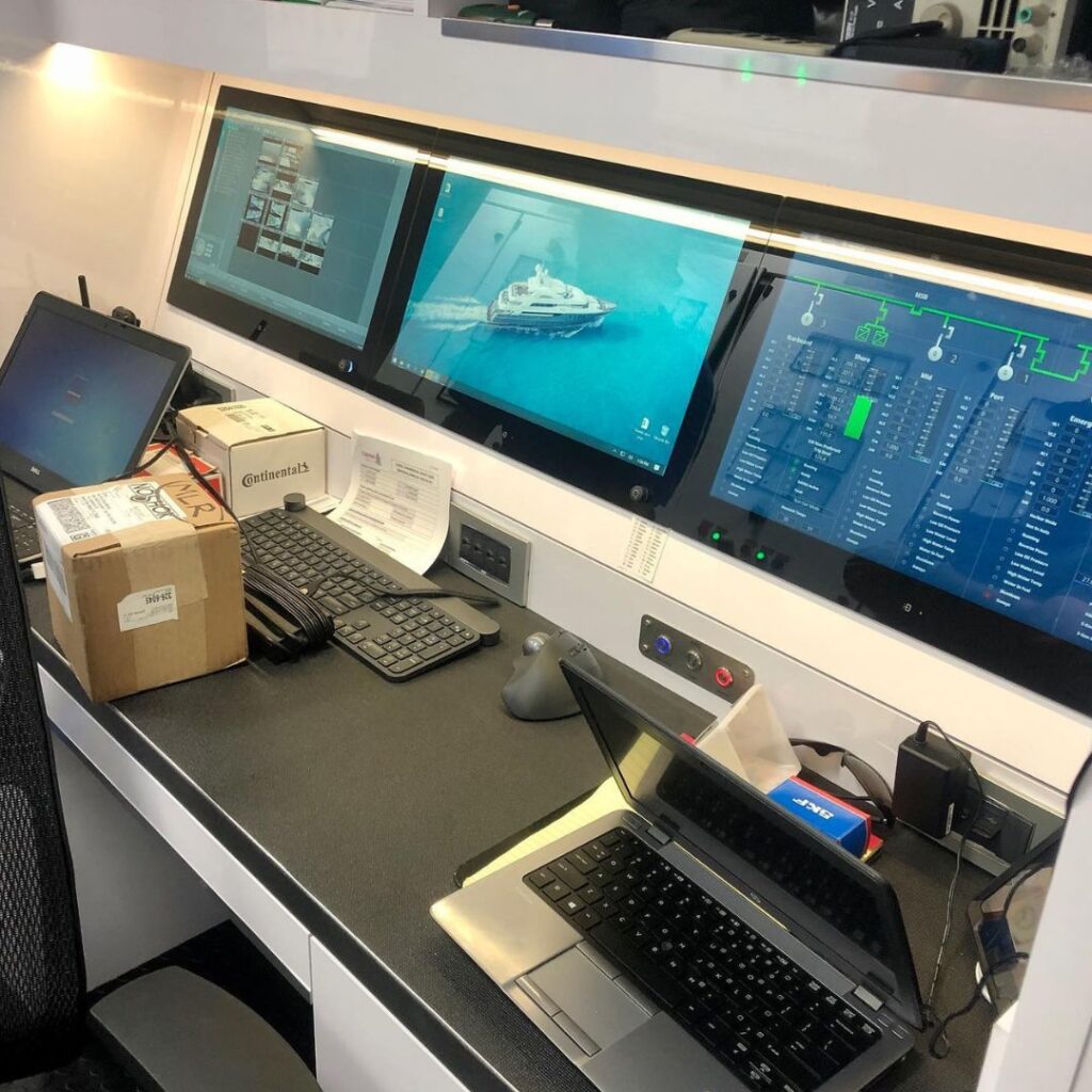 Marine power system control station with multiple screens displaying yacht monitoring software, a laptop, and a package, emphasizing the importance of efficient power management and upgrades in yacht maintenance.