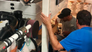 Two technicians working on a yacht's electrical system, examining components and making upgrades in a marine engine room.