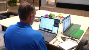 Person in blue shirt working at a table with two laptops, discussing marine electrical systems and documentation continuity during a roundtable meeting.